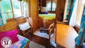 A cozy cabin interior with wooden walls, a bed with a yellow bedspread, a dining table, and a window with curtains. - Gîtes de France® - Roulotte de la Paille (LENDOU-EN-QUERCY)
