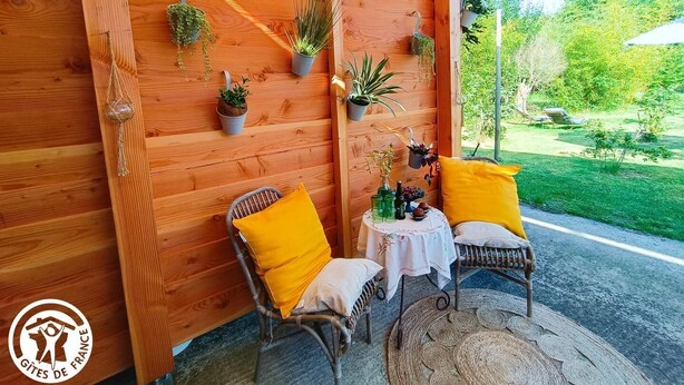 A wooden wall with potted plants, two wicker chairs with yellow cushions, and a small table with a white tablecloth. - Gîtes de France® - Roulotte de la Paille (LENDOU-EN-QUERCY)