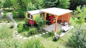 A small cabin with a covered porch and a red interior, surrounded by a lush garden and a stone pathway. - Gîtes de France® - Roulotte de la Paille (LENDOU-EN-QUERCY)