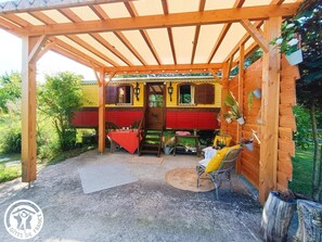 A wooden pergola with a red and yellow building underneath. There is a table with a red tablecloth and a chair with a yellow cushion. - Gîtes de France® - Roulotte de la Paille (LENDOU-EN-QUERCY)