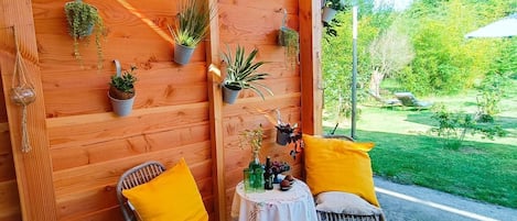 Una pared de madera con plantas en macetas, dos sillas de mimbre con cojines amarillos y una pequeña mesa con mantel blanco.