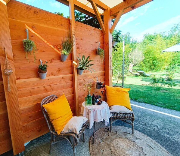 A wooden wall with potted plants, two wicker chairs with yellow cushions, and a small table with a white tablecloth.