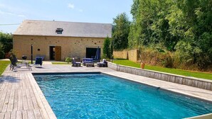 Una piscina rectangular con un deck de madera, rodeada de sillas de descanso y un edificio con techo inclinado al fondo.
