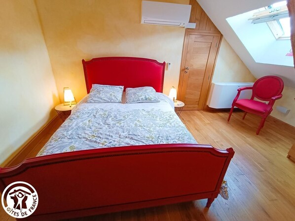 A bedroom with a red bed, two bedside lamps, a wooden wardrobe, a red chair, and a skylight.