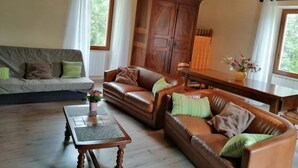 Ein gemütliches Wohnzimmer mit einer Couch, zwei Ledersesseln, einem Holztisch und einem Schrank aus Holz. An der Wand hängt eine Uhr, und von der Decke baumeln Lichterketten.
