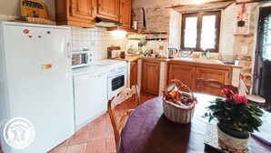 A rustic kitchen with wooden cabinets, a white refrigerator, a stove, and a microwave. There is a window with a view of the outside, and a basket with fruits on the table. - Gîtes de France® - Gîte in Negral (SAINT-VINCENT-DU-PENDIT)
