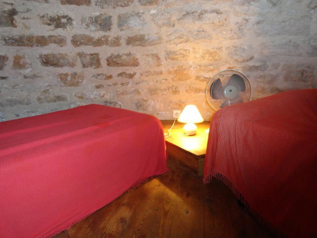 Una habitación con dos camas, suelo de madera, pared de ladrillo y una mesa con lámpara y ventilador.
