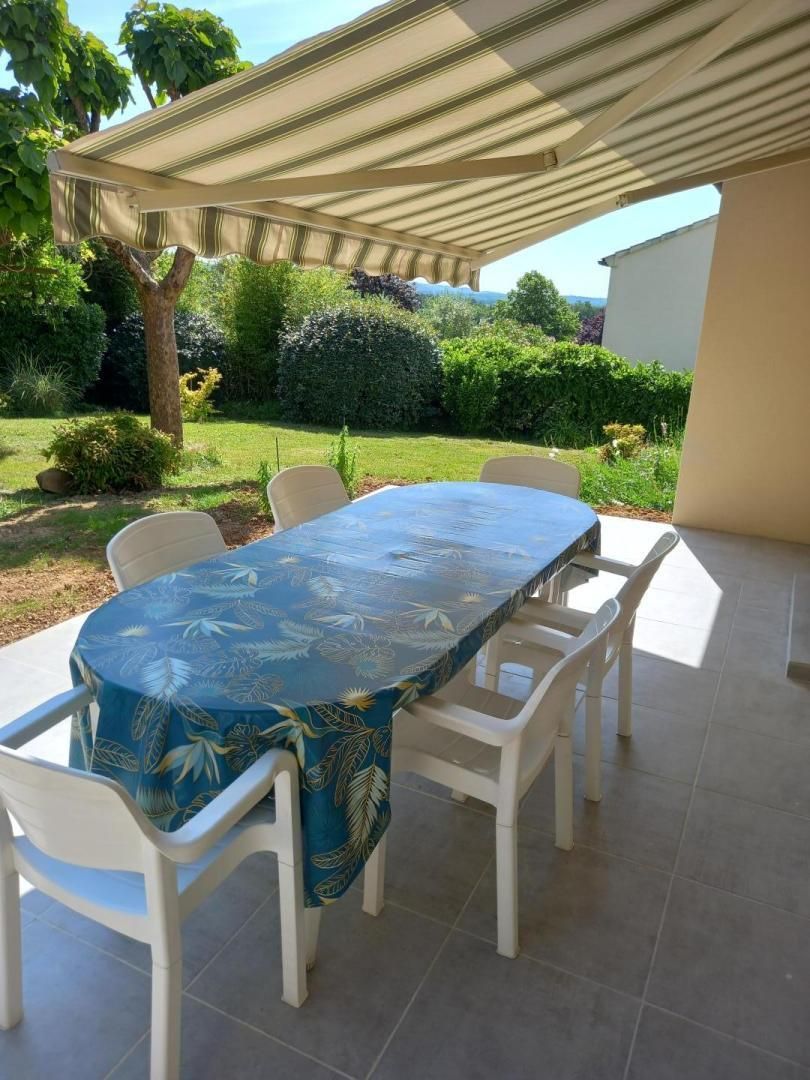 Een terras buiten met een tafel en stoelen, omgeven door een tuin en een overdekt gedeelte.