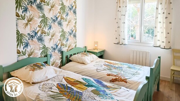 Ett sovrum med två sängar, ett fönster med gardiner, en sänglampa och en vägg med ett tropiskt bladmotiv.