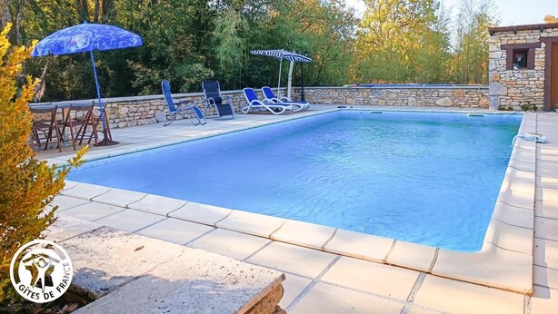 A swimming pool with lounge chairs and umbrellas, surrounded by stone walls and trees. - Gîtes de France® - Le Gîte des Buis du Causse (SAINT SULPICE)
