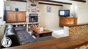 A cozy living room with a stone fireplace, wooden beams, a leather sofa, and a television. - Gîtes de France® - Le Gîte des Buis du Causse (SAINT SULPICE)