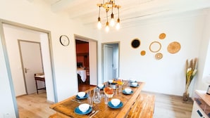 A dining area with a wooden table set for four, a clock on the wall, and a kitchen area visible in the background. - Gîtes de France® - La Vue Ombragée (VIAZAC)