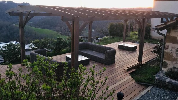 Una terraza de madera con mobiliario exterior, una pérgola y vistas a un paisaje montañoso.
