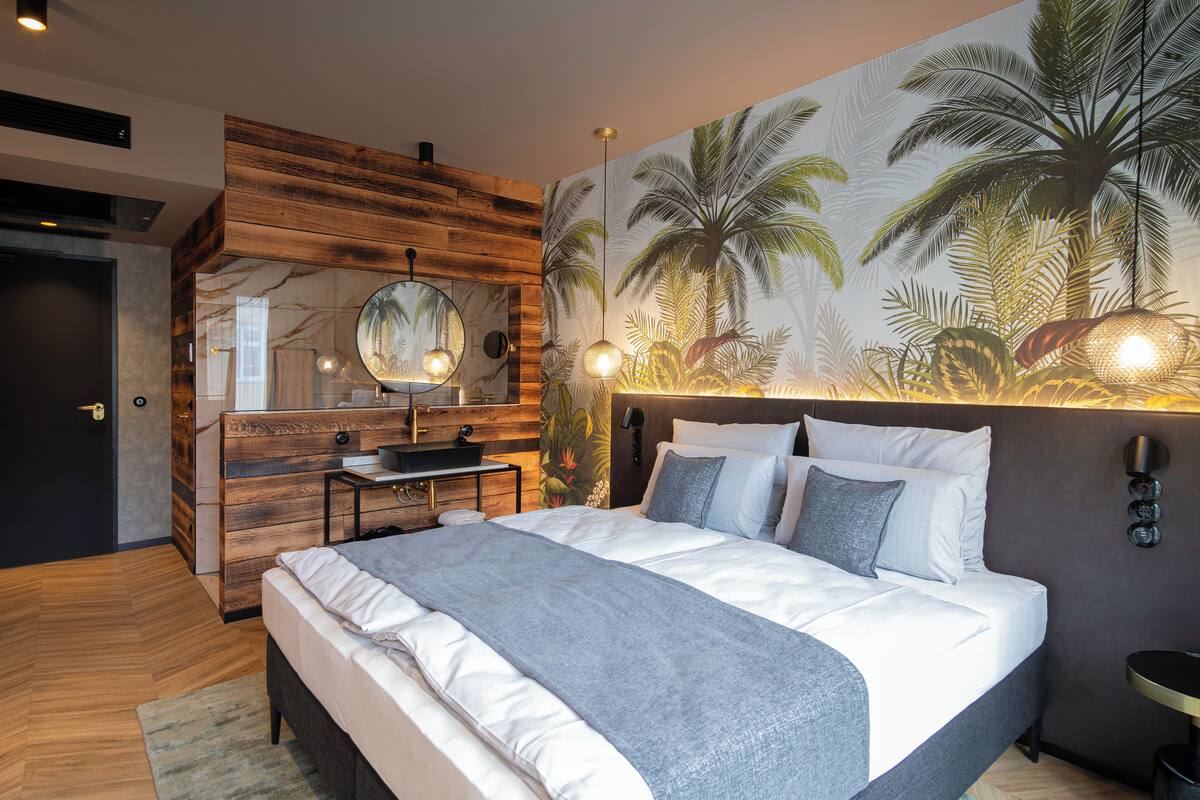 A modern bedroom with a wooden accent wall, a large bed with white bedding, a round mirror, and a wall mural of palm trees.