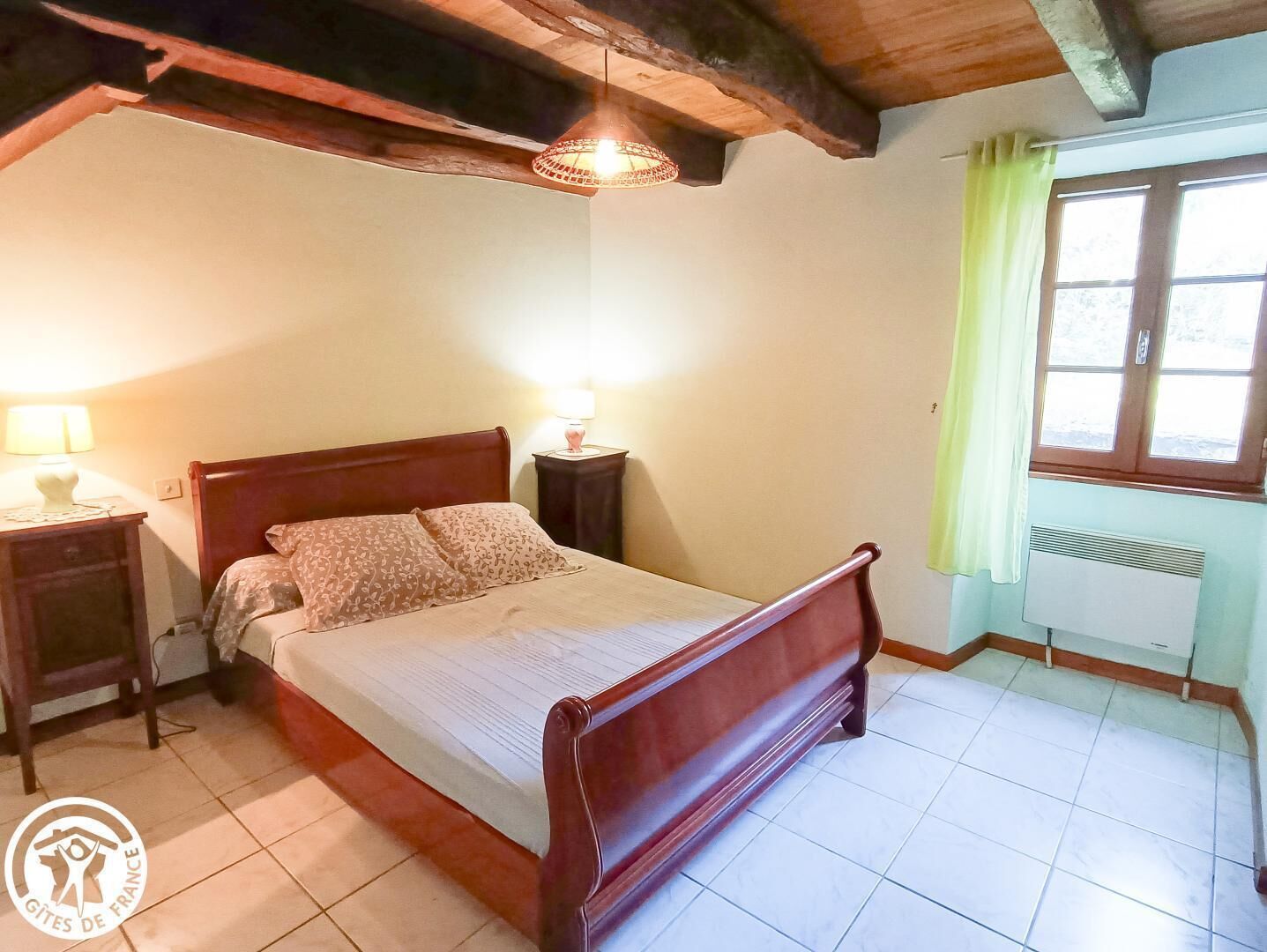 A bedroom with a wooden bed, two bedside tables with lamps, a window with a green curtain, and a radiator.
