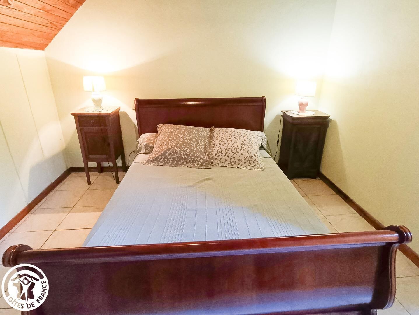 A bedroom with a wooden bed, two bedside tables with lamps, and a tiled floor.