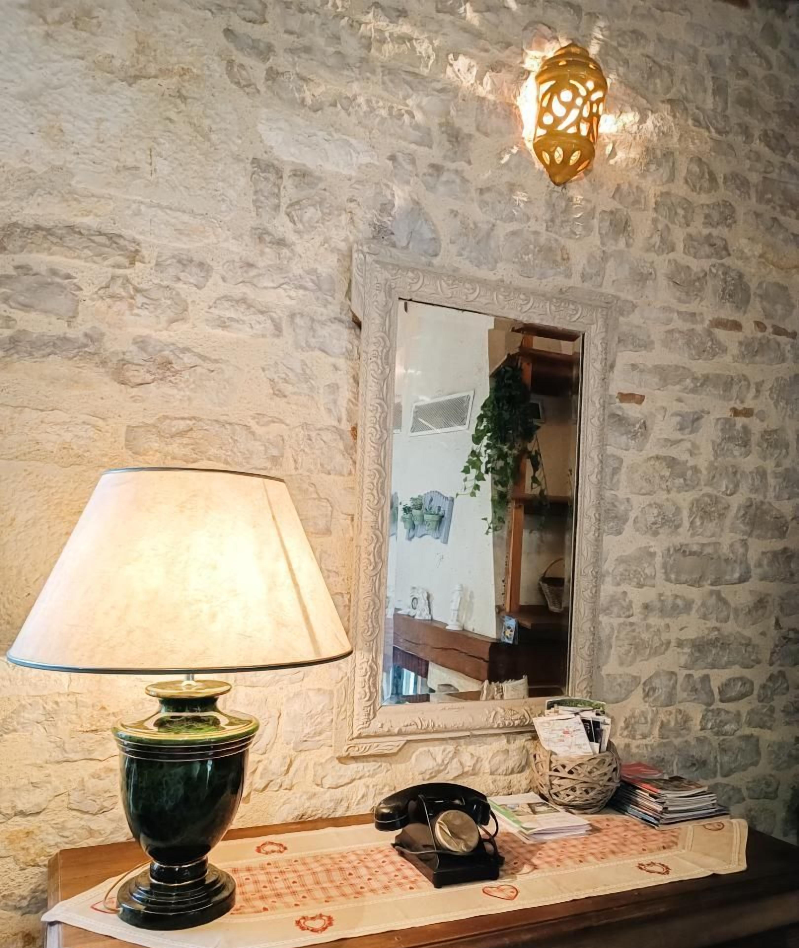 A stone wall with a mirror reflecting a plant and a shelf. A lamp, a telephone, and a basket with papers are on a wooden table.