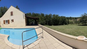 Una piscina con solárium empedrado, una casa con tejado inclinado y un césped con árboles al fondo.