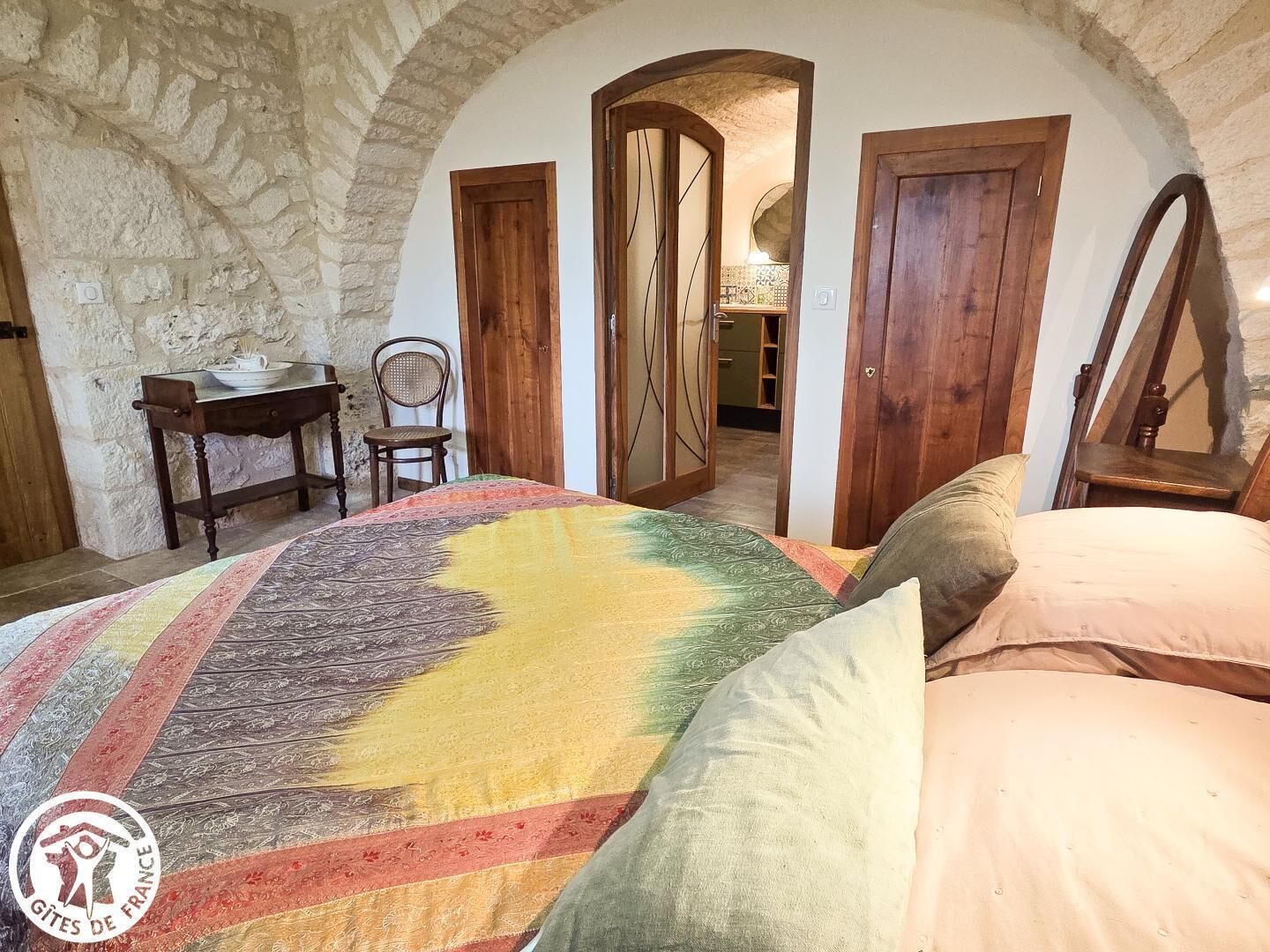 Een slaapkamer met een stenen muur, houten deuren, een bed met een kleurrijk dekbed, een stoel en een klein tafeltje met een schaal.