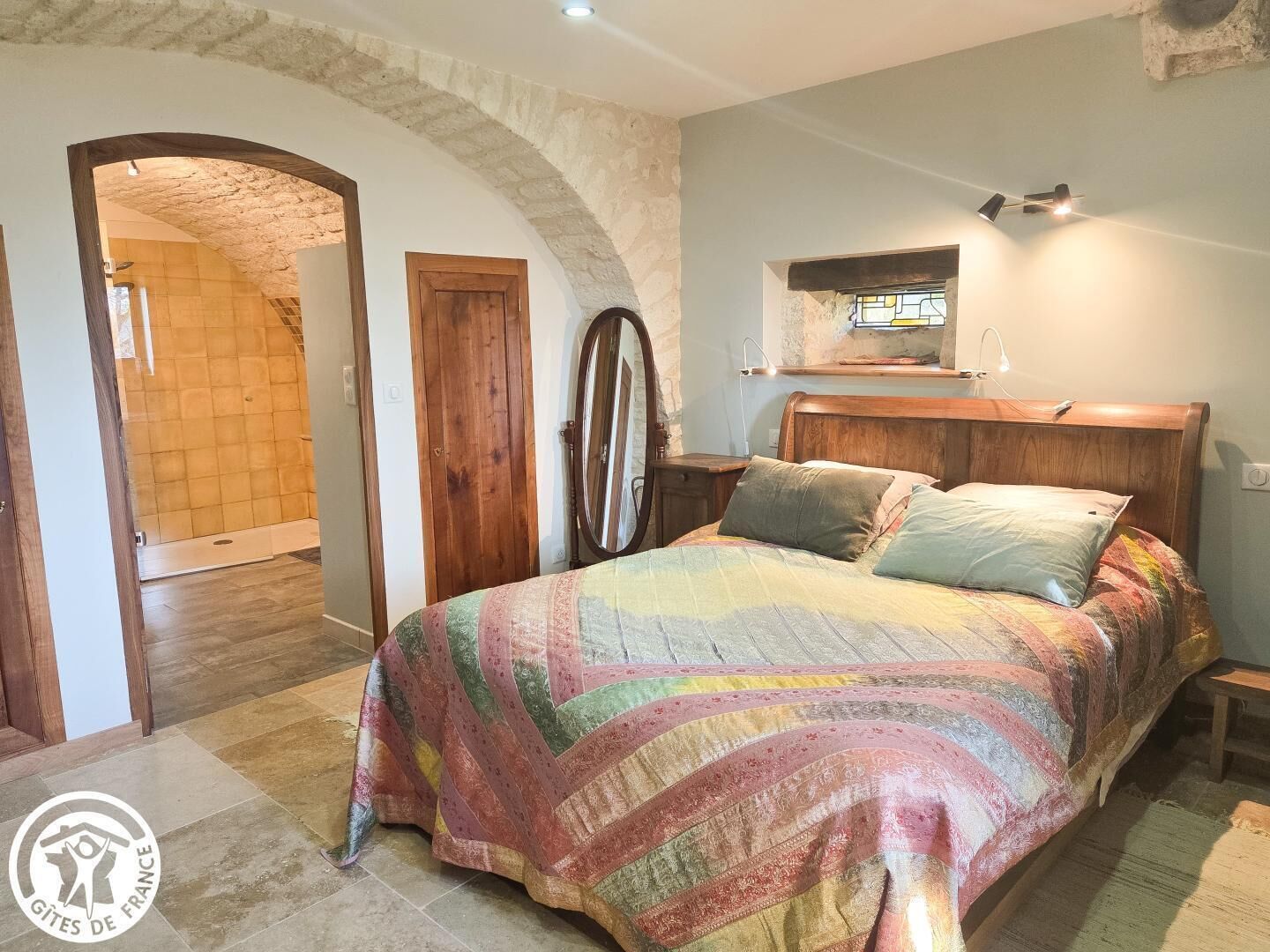 Un dormitorio con cama, cabecera de madera, mesita de noche, espejo y ventana con vistas al exterior.