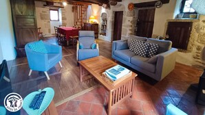 A cozy living room with a wooden coffee table, blue armchairs, and a grey sofa. There is a dining area with a red table and chairs, and a stone fireplace. - Gîtes de France® - GITE LA BUISSIERE (GREALOU)