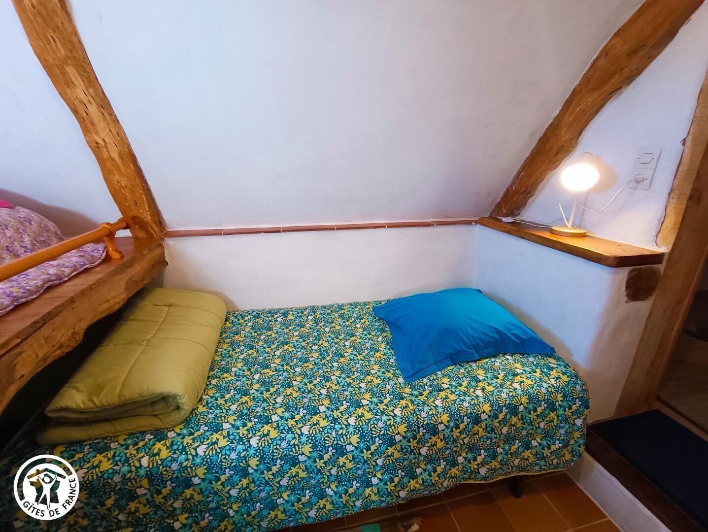 A small, narrow room with a single bed, a wooden shelf, and a desk with a lamp.