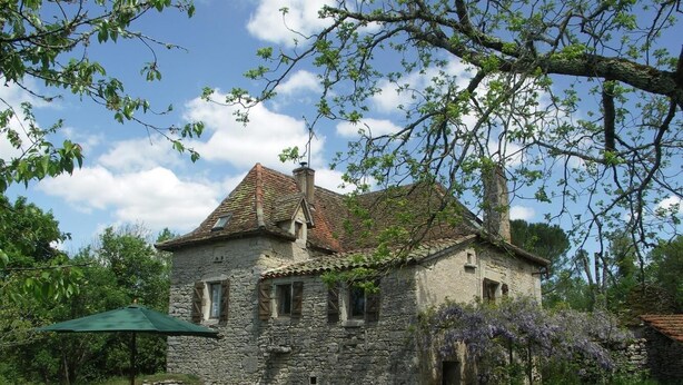 A stone house with a tiled roof, a small patio with a table and chairs, and a lush garden with purple flowers. - Gîtes de France® - GITE LA BUISSIERE (GREALOU)