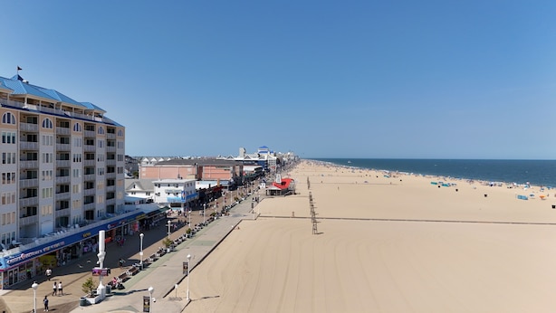 Un hôtel en bord de mer, avec plusieurs étages, une promenade bordée de boutiques et, en arrière-plan, une plage de sable.