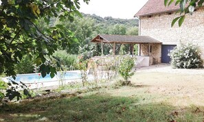 A stone building with a tiled roof, a swimming pool, and a covered outdoor area with plants and a chair.