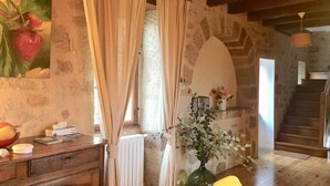 A rustic room with a stone wall, wooden furniture, a white radiator, a vase with flowers, and a staircase in the background.