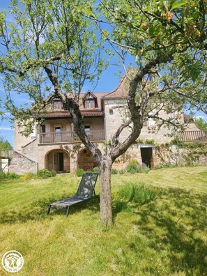 A stone house with a balcony, a tree in the foreground, and a grassy lawn.
