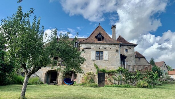 A stone house with a pitched roof, a visible chimney, and a small garden area with a tree and a blue pool.