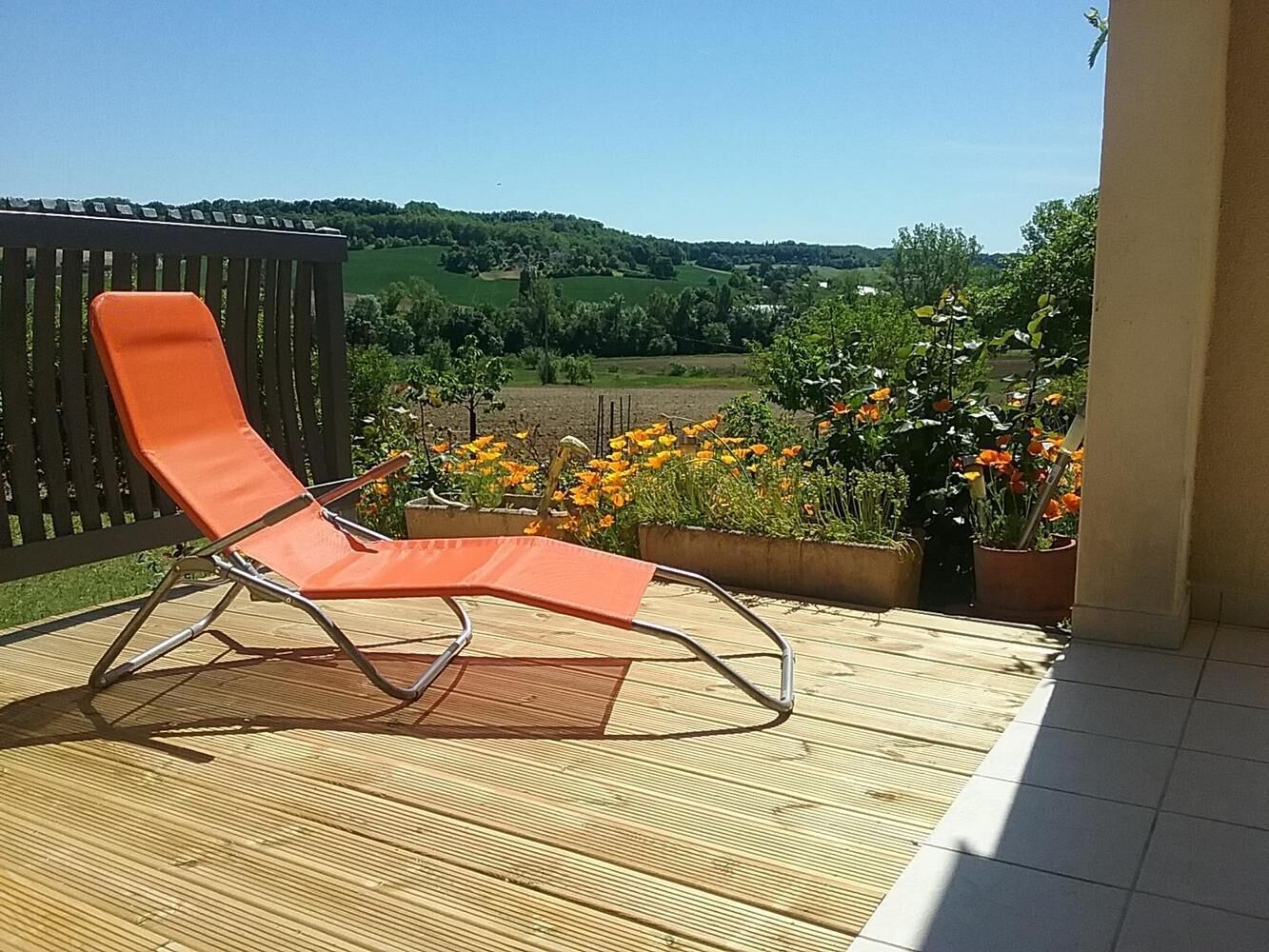 A wooden deck with a metal-framed orange chair, potted plants, and a view of a green landscape.