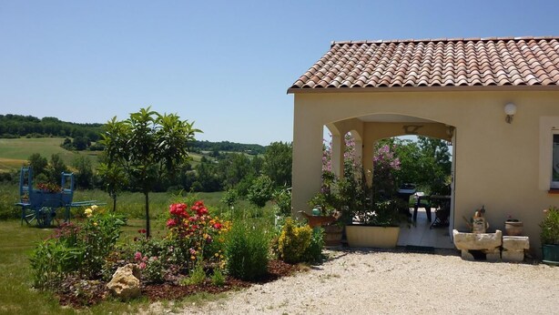 A building with a tiled roof, a gravel driveway, and a garden with various plants and flowers. - Gîtes de France® - Jane's house (FLAUGNAC)