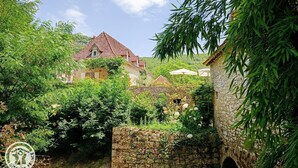 Um edifício de pedra tradicional com telhado de palha, rodeado por vegetação exuberante e um céu azul límpido.