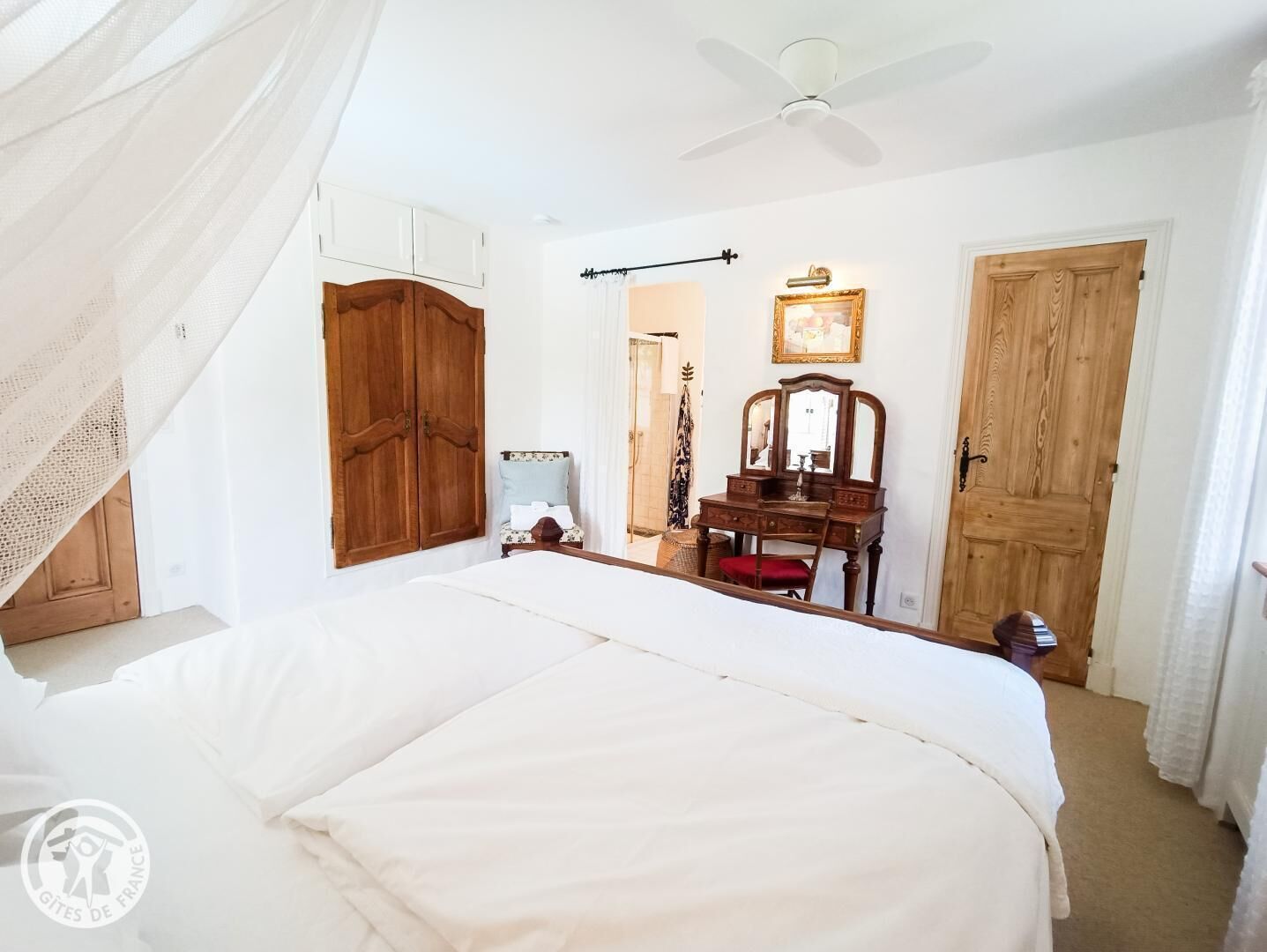 A bedroom with a large bed, wooden wardrobe, a vanity with a mirror, and a ceiling fan.