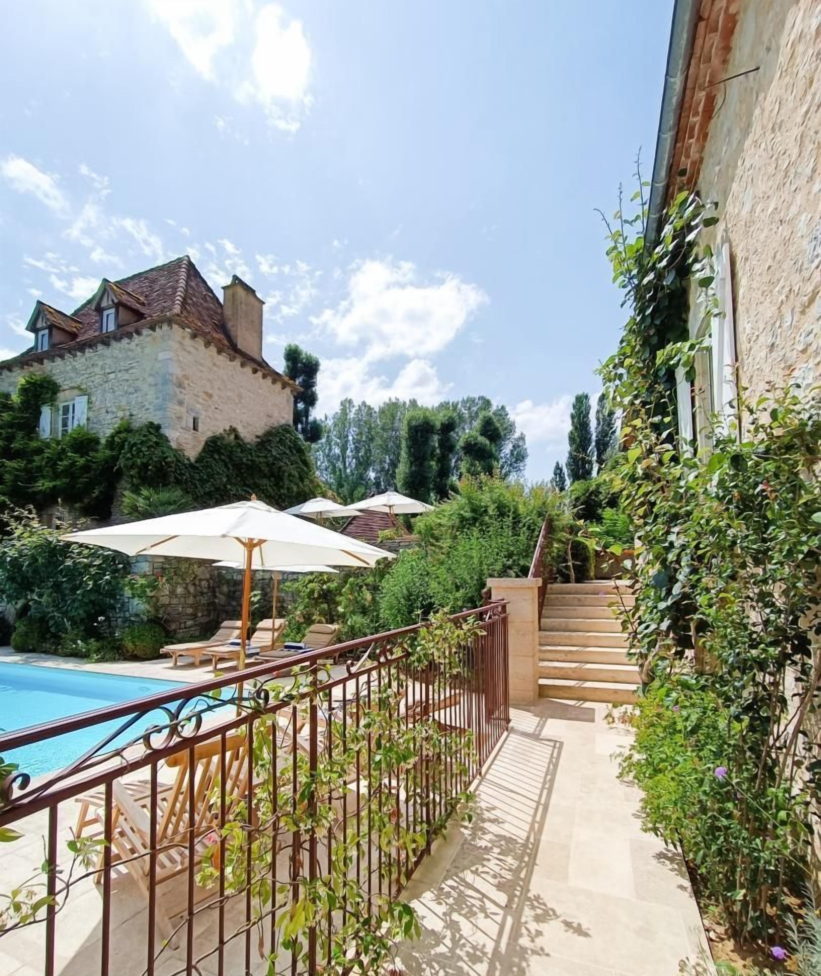 Ein Poolbereich mit Liegestühlen und Sonnenschirmen, im Hintergrund ein Steinhaus und eine Treppe, die zu einer höheren Ebene führt.