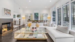 A modern living room with a glass coffee table, white sofas, a fireplace, and large windows.
