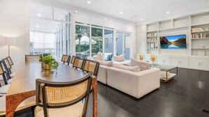 A spacious living room with a large dining table, comfortable seating, a sofa, and a television on a white wall.
