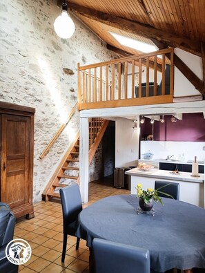 Un interior rústico con una escalera de madera que conduce a un altillo, un comedor con mesa y sillas, y una zona de cocina con fregadero y encimera.