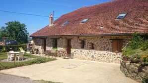 Um edifício de pedra com telhado em tijolos, um pátio com móveis de exterior e uma pequena área de jardim.