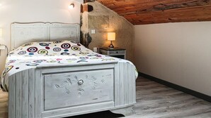 Quarto com teto de madeira, cama com colcha colorida, mesa de cabeceira com abajur e uma pequena janela.