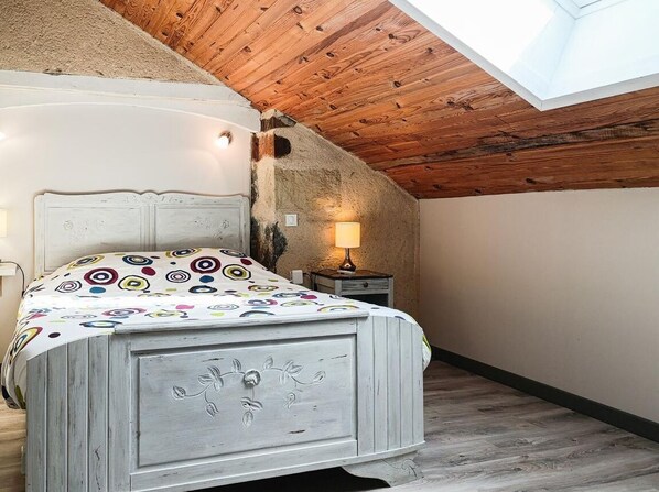 Un dormitorio con techo de madera, una cama con colcha colorida, una mesita de noche con lámpara y un ventanal pequeño.