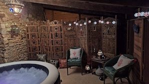 Una habitación acogedora con pared de piedra, paneles de madera y un amplio jacuzzi. Hay dos sillones de mimbre y una mesita con una lámpara.