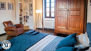 Una habitación acogedora con una cama, un sillón de cuero marrón, un armario de madera, una estantería blanca con objetos decorativos y una ventana con luz natural.