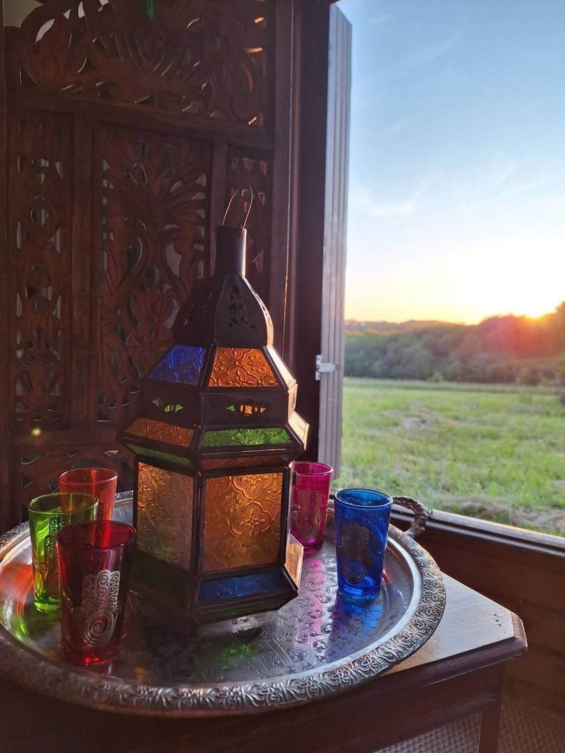 A traditional lantern with colorful glass panes, surrounded by matching glass cups on a tray, set against a backdrop of a wooden carved door and a view of a green landscape through a window.