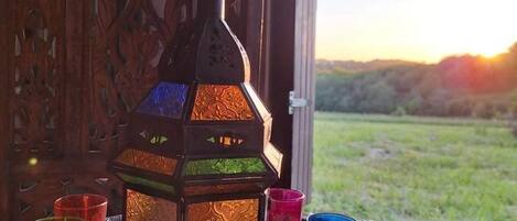 A traditional lantern with colorful glass panes, surrounded by matching glass cups on a tray, set against a backdrop of a wooden carved door and a view of a green landscape through a window.