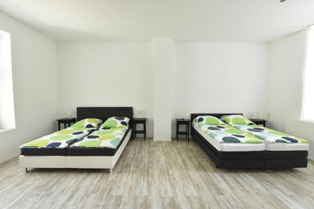 A room with two beds, each with a black headboard and a green and white patterned bedspread. There are two bedside tables with lamps, and the room has a light wooden floor.