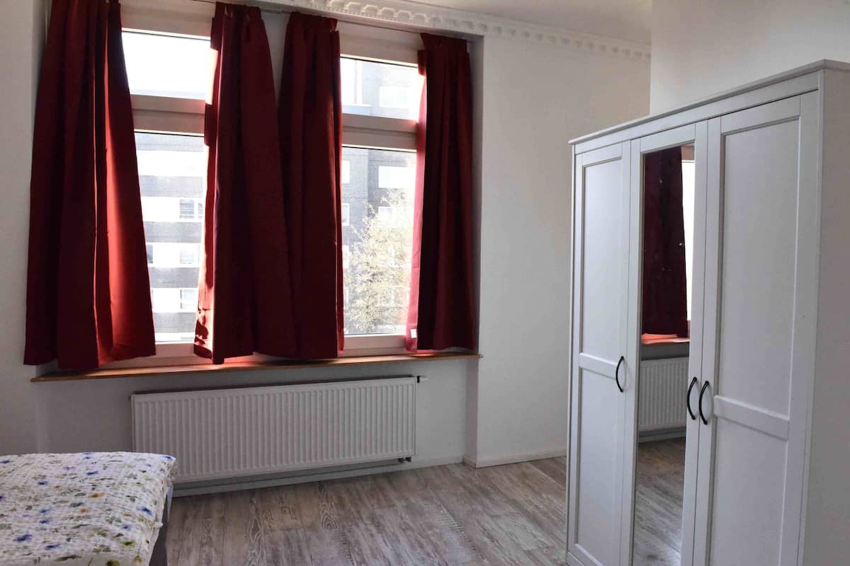 A bedroom with a bed, a window with red curtains, a radiator, and a wardrobe.