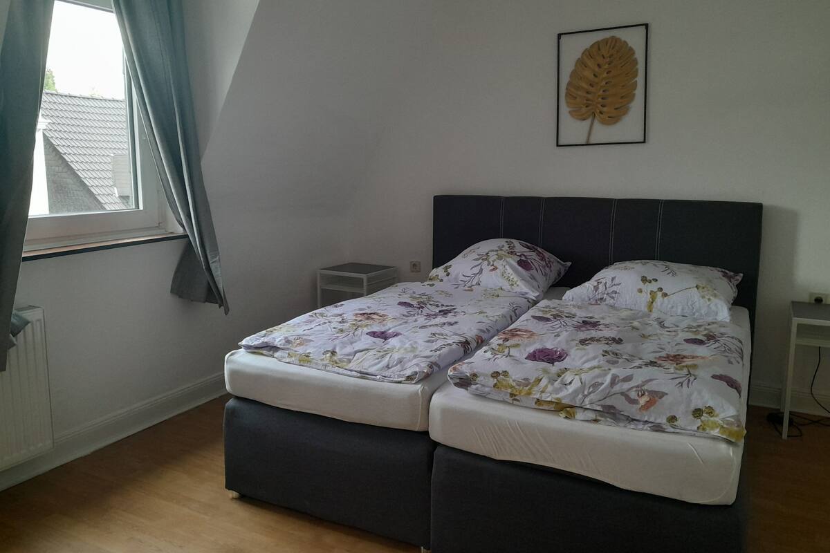 A bedroom with two beds, a small table, a radiator, and a window with curtains.
